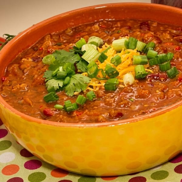 Hearty chili in a yellow bowl, topped with shredded cheese, green onions, and cilantro, with fresh tomatoes and garlic.