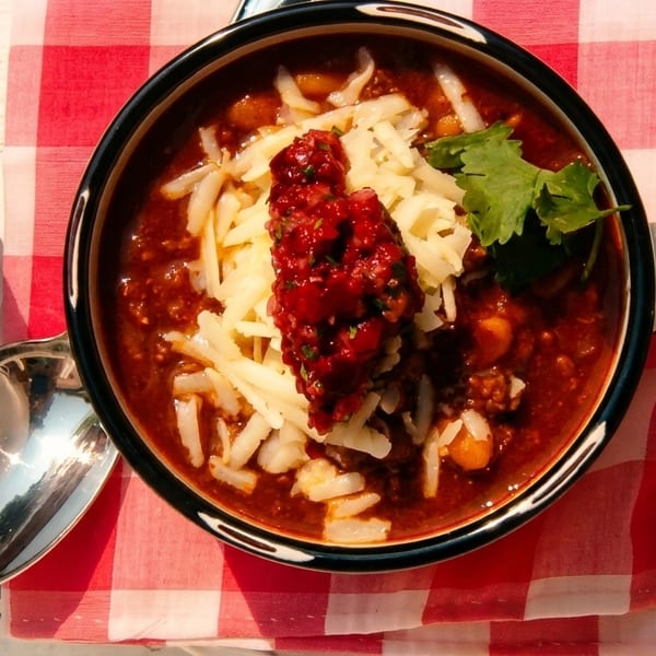 Overhead view of a bowl of chili with shredded cheese, salsa, and cilantro on a red and white checkered napkin.